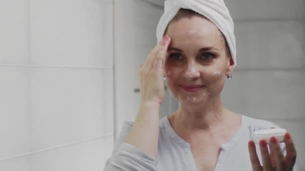 Volwassen vrouw met een handdoek op haar hoofd aanbrengen crème en kijken in de spiegel genieten van natuurlijke schoonheid en gezond — Stockvideo