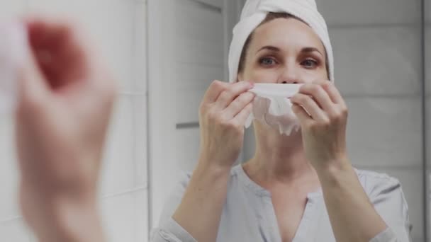 Взрослая женщина с полотенцем на голове снимает маску на лице, глядя в зеркало — стоковое видео