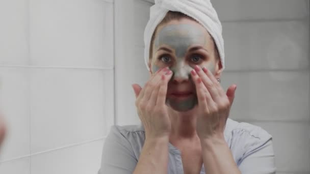 Mujer adulta con una toalla en la cabeza aplicando máscara de arcilla en su cara mirando a la cámara — Vídeo de stock