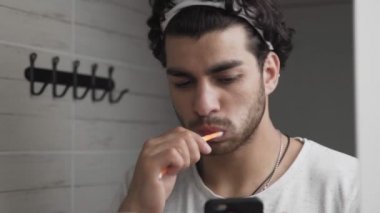 Sakallı adam dişlerini fırçalıyor. Banyoda telefona bakıyor.