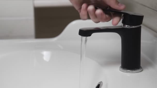 Männliche Hand dreht schwarzen Wasserhahn zum Gießen von Wasser in Waschbecken im Badezimmer. Wasser läuft aus Wasserhahn. — Stockvideo