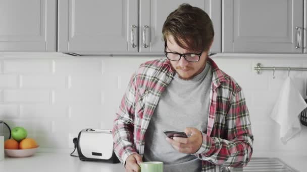 Jovem usando telefone e beber café enquanto sandng na cozinha — Vídeo de Stock