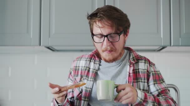 Young man eating toast with chocolate paste and drinking coffee or tea — Stock Video