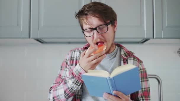 Young man eating toast with strawberry jam and reading a book — Stock Video