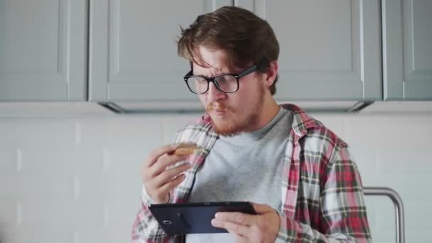 Jovem com tablet comendo torrada com pasta de chocolate. Homem assistir a um vídeo engraçado no tablet e rir — Vídeo de Stock