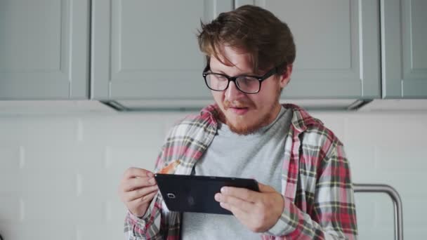 Молодой человек с планшетом ест тосты с клубничным джемом. Мужчина смотрит смешное видео на планшете и смеется — стоковое видео