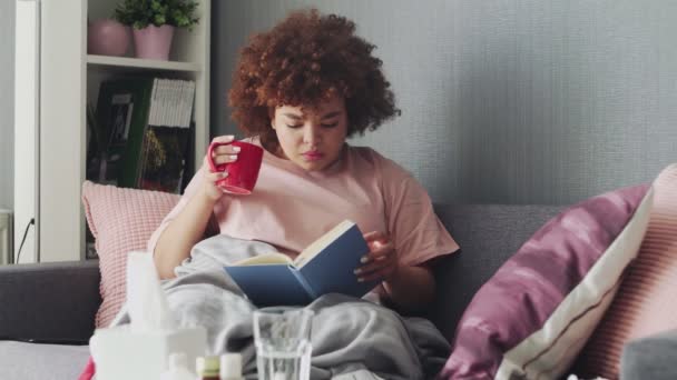 一个女人在家里感冒了。躺在沙发上，拿着一本书和一杯热茶，上面铺着一条毯子 — 图库视频影像