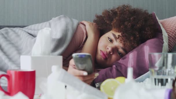 Doente menina americana africana com gripe em casa, jovem mulher negra doente com febre assistindo programa de tv e usando controle remoto . — Vídeo de Stock