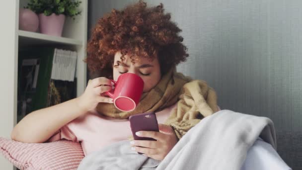 Doente Jovem afro-americana usando telefone e bebendo um chá quente em casa — Vídeo de Stock