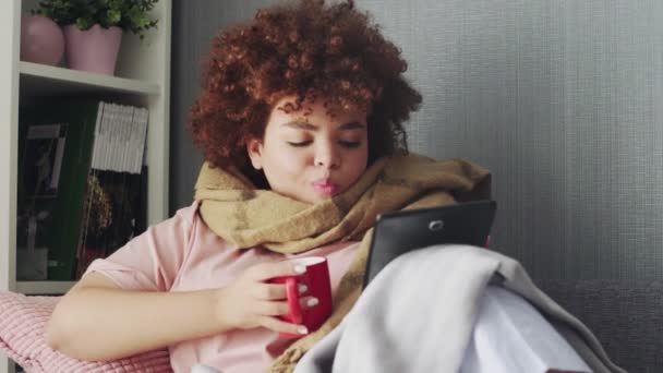 Doente Jovem afro-americana usando tablet pc e beber um chá quente em casa — Vídeo de Stock