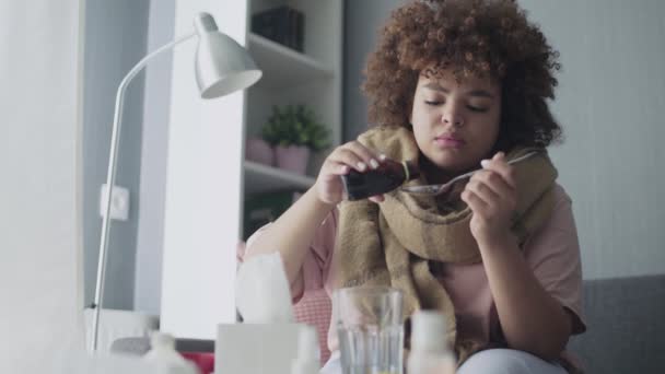 Fille afro-américaine malade verser des médicaments ou du sirop antipyrétique à la cuillère et de le boire avec de l'eau, assis sur le canapé . — Video