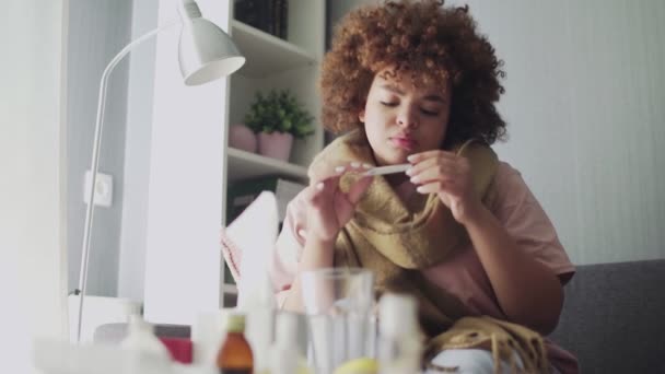 Beteg afro-amerikai lány influenzával, aki otthon fekszik a kanapén. I ll fiatal fekete nő hideg ellenőrzése hőmérséklet hőmérővel — Stock videók