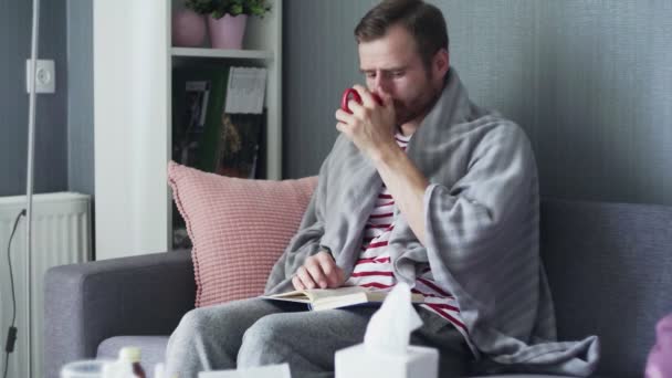 Um homem adulto doente senta-se no sofá coberto com xadrez cinza, lê um livro e bebe um chá quente. — Vídeo de Stock