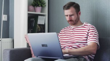 Hüsrana uğramış ve yorgun genç adam dizüstü bilgisayarda çalışıyor. Kucağında tutuyor ve evdeki bir koltukta oturuyor..