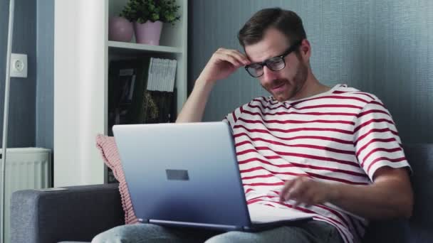 Κουρασμένος νεαρός άνδρας με γυαλιά εργάζεται σε ένα φορητό υπολογιστή κρατώντας το στο Lap του και κάθεται σε έναν καναπέ στο σπίτι. — Αρχείο Βίντεο