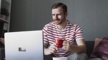 Sakallı, gülümseyen adam çay içiyor ve bilgisayarı kaydırıyor..