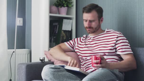 Κάθισμα νεαρού άνδρα στον καναπέ στο σαλόνι και να διαβάσετε το βιβλίο με τσάι ή καφέ κύπελλο — Αρχείο Βίντεο