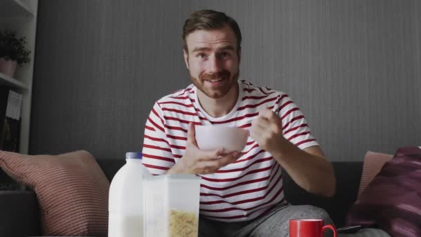 Joven viendo divertido programa de televisión durante el desayuno sentado en el sofá en la habitación — Vídeos de Stock