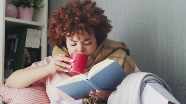 Una mujer tiene un resfriado en casa. Acostado en el sofá con un libro y una taza de té caliente, cubierto con una manta — Vídeos de Stock