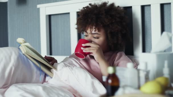 La chica afroamericana enferma tiene un resfriado en casa. Acostado en la cama con un libro y una taza de té caliente — Vídeos de Stock