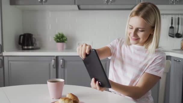 Çekici kadın, mutfakta kahvaltı ederken tablet üzerinde video oyunu oynuyor. — Stok video