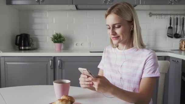 Aantrekkelijke vrouw met hoofdtelefoon, zittend aan tafel, luisterend naar muziek in haar smartphone en dansend. — Stockvideo