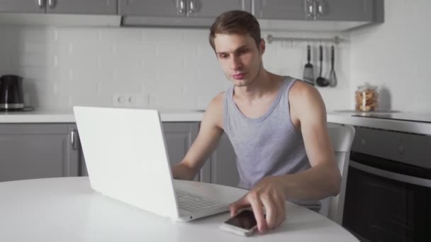 若い男はノートパソコンや自宅で携帯電話を使用して作業します。忙しいビジネスマンだ — ストック動画