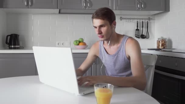 Joven freelancer con auriculares trabajando en computadora portátil y bebiendo jugo — Vídeos de Stock