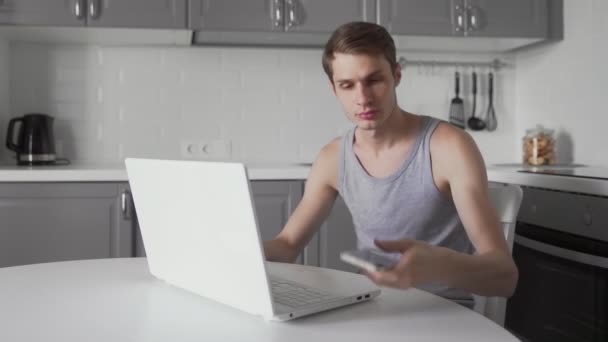 Молодой предприниматель разговаривает по мобильному телефону и работает над ноутбуком на кухне . — стоковое видео