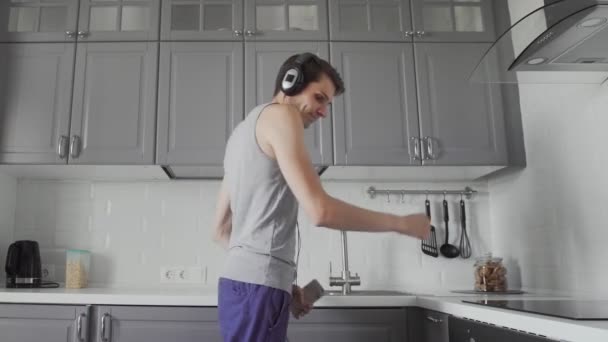 Jóképű fiatal vicces férfi fülhallgatóban táncol a konyhában reggel, és érezd jól magad a nyaraláson. — Stock videók