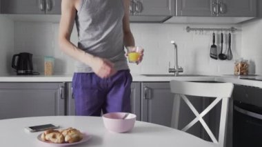 Genç adam mutfakta kahvaltı yapıyor. Adam meyve suyu içiyor ve telefon kullanıyor.