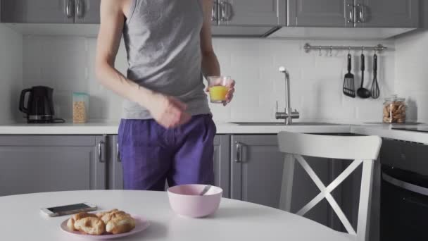 Młody człowiek je śniadanie w kuchni. Mężczyzna pije sok i używa telefonu — Wideo stockowe