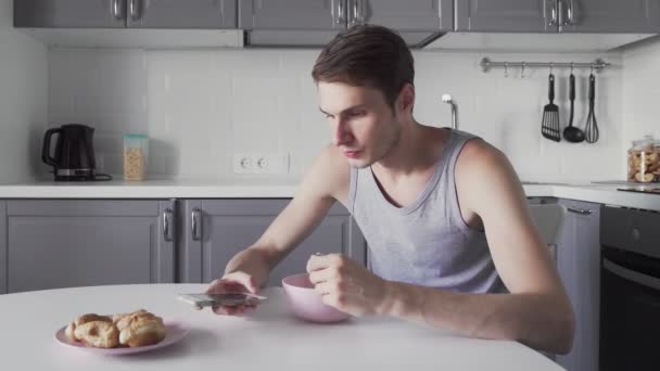 若い男はスープを食べ、台所で電話を使用して — ストック動画