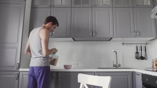 Jovem preparando café da manhã na cozinha. ele deita flocos de milho numa tigela — Vídeo de Stock