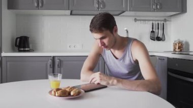 Genç adam evde mutfakta otururken dijital tablet kullanıyor.