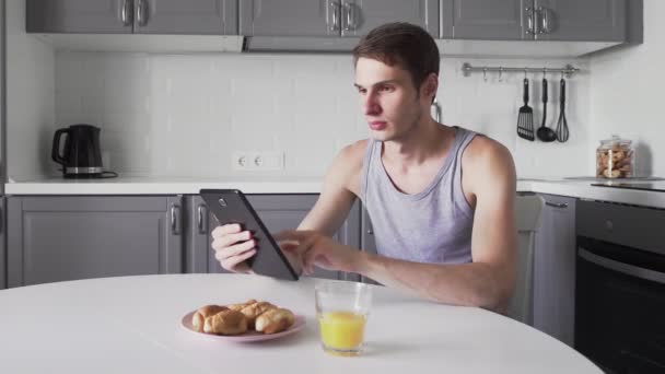 Hombre charlando en la computadora de la tableta y beber jugo por mesa en la cocina — Vídeo de stock