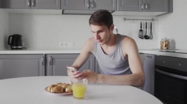 Genç adam portakal suyu içiyor ve elinde akıllı bir cihaz kullanıyor. Sabahları modern mutfakta oturan yakışıklı adamın günlük rutini.