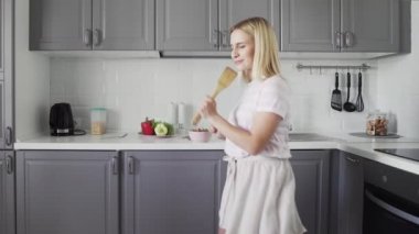 Ahşap bir spatula mikrofonu tutan, karaoke söyleyen, dans eden, mutfakta yemek yapan mutlu, neşeli, neşeli genç bir kadın.