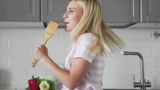 Close up of happy active funny young woman holding wooden spatula microphone singing karaoke song dancing listening music cooking in kitchen — Stock Video