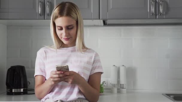 El primer plano de la muchacha joven por la mañana en la cocina que comprueba teléfono. Mujer charlando con amigos usando un teléfono móvil. Chica escribiendo un mensaje en el teléfono inteligente . — Vídeos de Stock