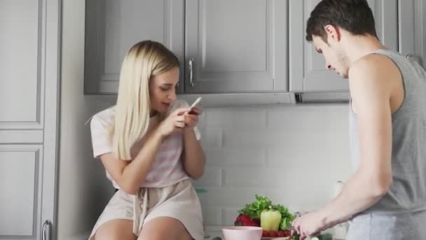 Egy lány lefényképez egy fickót, aki salátát szeletel ebédre a konyhában. — Stock videók