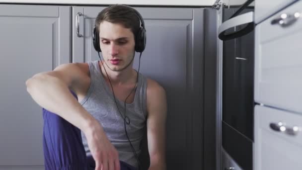 台所の床に座ってヘッドフォンを身に着けているスマートフォンの音楽を聞いている若い男 — ストック動画