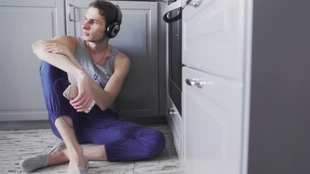 Jeune homme écoutant de la musique sur smartphone dans les écouteurs, dansant, assis sur le sol dans la cuisine et regardant par la fenêtre — Video