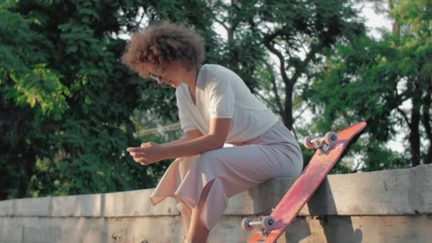 Retrato de una joven hipster con un monopatín y teléfono sentado en el parque — Vídeo de stock