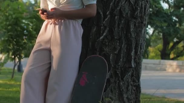 Кучерява жінка зі скейтбордом використовує мобільний телефон у громадському парку . — стокове відео