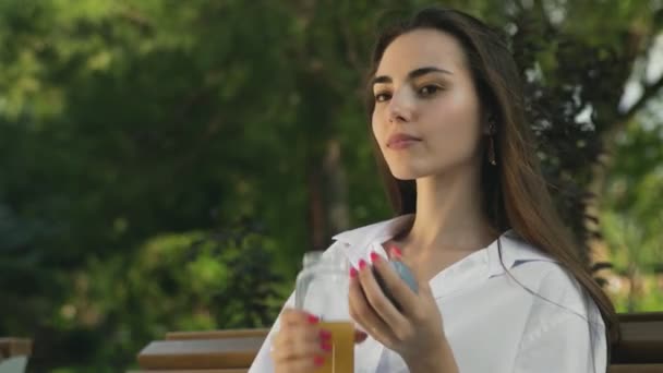 在夏季公园，年轻漂亮的姑娘在户外喝着瓶子里的橙汁. — 图库视频影像