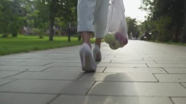Mavi pantolonlu ve beyaz ayakkabılı genç bir kadının arka manzarasında pamuk örgüsü ile meyve dolu alışveriş torbaları ve yaz parkında yürüyüşler var. Sıfır atık.