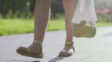 Kıllı bacaklı genç bir kadın, elinde sebzelerle pamuk örgüsü alışveriş torbası tutuyor ve yaz parkında yürüyor. Kadın gücü, vücut pozitifliği