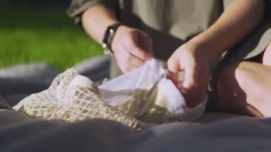 Genç bir kadın battaniyeye oturur ve yaz parkında pamuk örgüsü alışveriş çantasından elma alır. Sıfır atık.