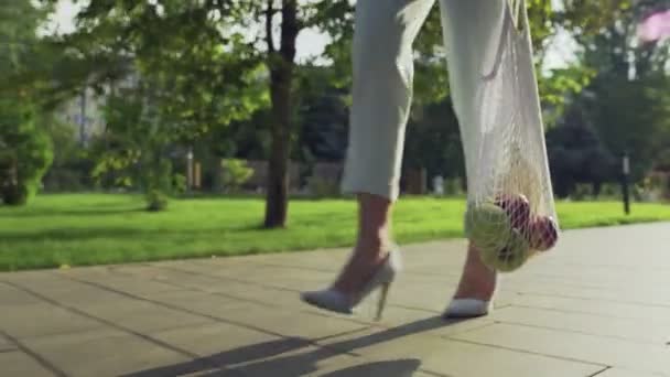 Felismerhetetlen nő tartja pamut háló bevásárló táska gyümölcsökkel és séták a nyári parkban — Stock videók